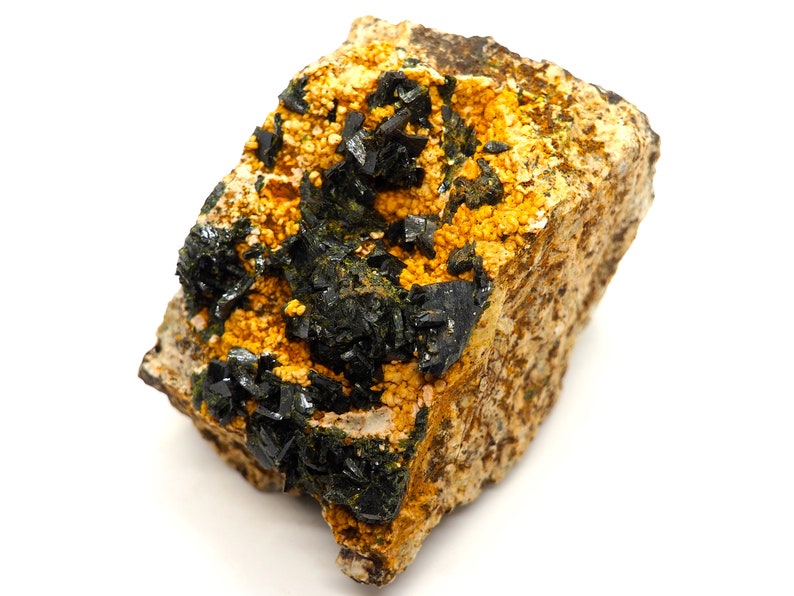 May include: A close-up of a mineral specimen featuring a rough, tan-colored matrix. The matrix is speckled with small, golden-yellow crystals and clusters of dark, black, crystalline structures. The mineral has a natural, earthy appearance.