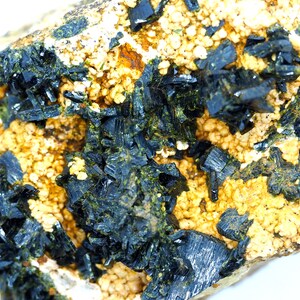 May include: Close-up of a mineral specimen featuring dark, crystalline structures clustered on a matrix of beige and yellow granular material. The crystals have a metallic sheen, and the overall texture is rough and uneven.