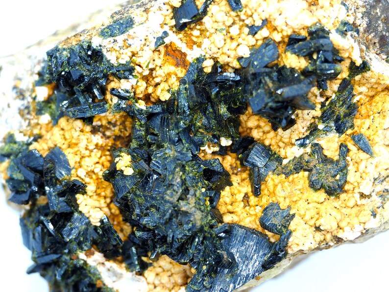 May include: Close-up of a mineral specimen featuring dark, crystalline structures clustered on a matrix of beige and yellow granular material. The crystals have a metallic sheen, and the overall texture is rough and uneven.