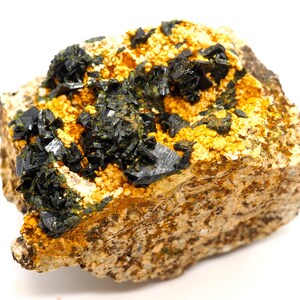 May include: A close-up of a mineral specimen featuring dark, crystalline formations atop a matrix of tan and yellow granular material. The mineral crystals are a deep, reflective black, contrasting with the lighter background.