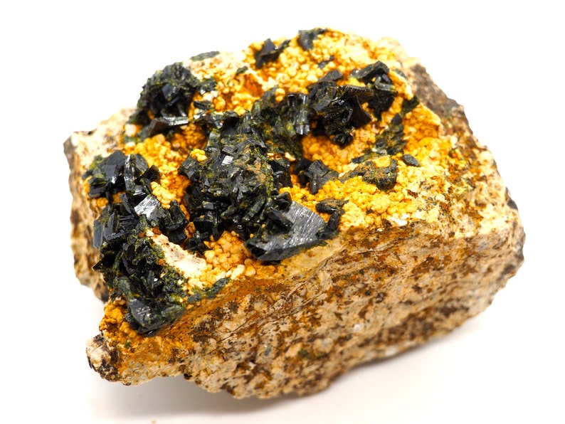 May include: A close-up of a mineral specimen featuring dark, crystalline formations atop a matrix of tan and yellow granular material. The mineral crystals are a deep, reflective black, contrasting with the lighter background.