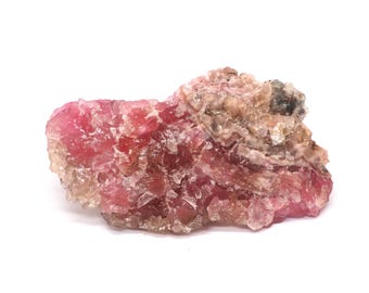 May include: A raw, uncut mineral specimen with a mix of pink, red, and tan hues. The crystalline structure is visible, with a rough, textured surface. The stone is set against a white background.
