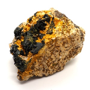 May include: A close-up of a mineral specimen featuring a rough, tan-colored rock matrix. Dark, crystalline structures and clusters of small, yellow-orange crystals are visible on the surface. The mineral has a natural, earthy appearance.