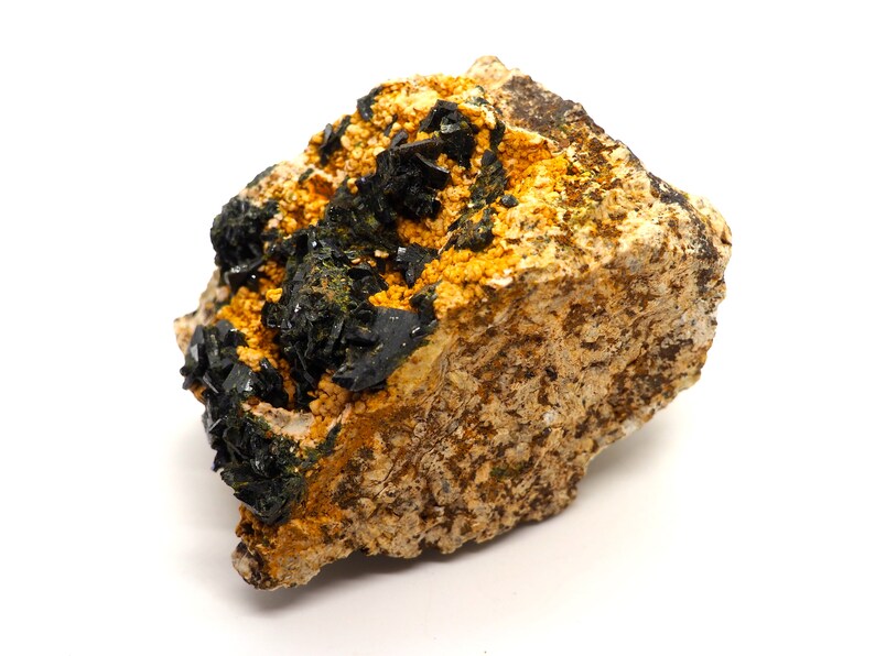 May include: A close-up of a mineral specimen featuring a rough, tan-colored rock matrix. Dark, crystalline structures and clusters of small, yellow-orange crystals are visible on the surface. The mineral has a natural, earthy appearance.