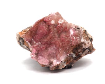May include: A natural mineral specimen featuring a cluster of pink and red crystalline formations. The rock has a rough, textured surface with areas of brown and tan hues, set against a white background.