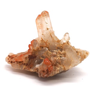 Peut inclure: Un groupe de cristaux de quartz transparents avec des inclusions rouges, enchâssés dans une matrice de roche brune et blanche. Les cristaux sont pointus et acérés, et la roche est rugueuse et texturée.