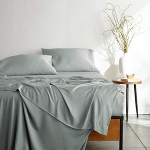 May include: A sage green bed with a white headboard and two pillows. The bed is made with a sage green sheet set and a matching duvet cover. The bed is in a bedroom with a white wall and a vase of branches on a side table.