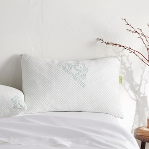 May include: A white pillow with a green and white pattern and the text "Bamboo is Better". The pillow is on a white bed with a white sheet.