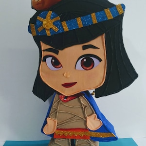 May include: A cartoon character pinata with a blue cape and a gold crown. The character has brown hair, brown eyes, and a red mouth. The pinata is made of cardboard and is decorated with glitter.