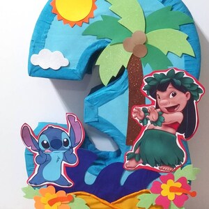 Puede incluir: Una pi&ntilde;ata azul n&uacute;mero tres con un tema tropical. La pi&ntilde;ata presenta a Stitch de Lilo & Stitch, Lilo, una palmera, un sol amarillo y flores.