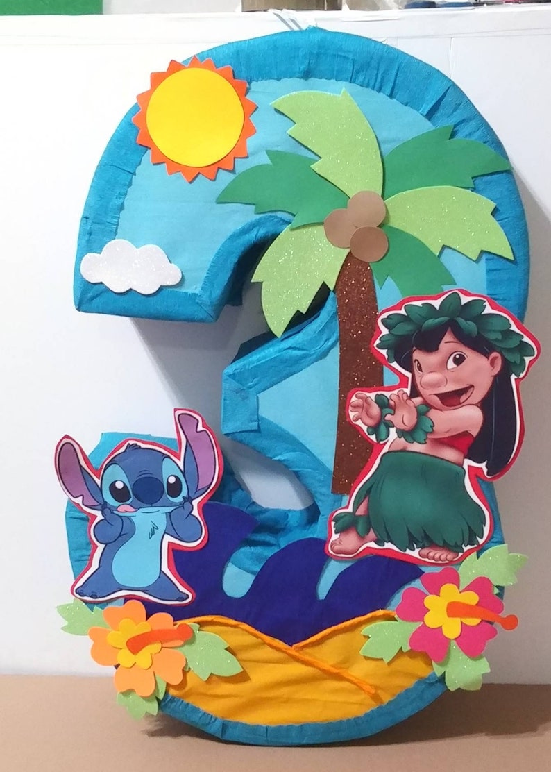 Puede incluir: Una pi&ntilde;ata azul n&uacute;mero tres con un tema tropical. La pi&ntilde;ata presenta a Stitch de Lilo & Stitch, Lilo, una palmera, un sol amarillo y flores.