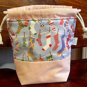 May include: A drawstring bag with a light blue patterned fabric featuring colorful socks hanging on a clothesline. The bottom and top trim are a solid light pink. The bag has a drawstring closure with cream-colored cords.