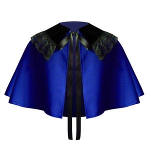 May include: A royal blue capelet featuring a black velvet collar with black lace trim along the edges. The garment includes a black satin ribbon tie at the center front neckline, allowing for closure. The fabric has a smooth, slightly reflective finish.