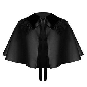 May include: A black capelet featuring a smooth satin-like fabric, a wide collar with black lace trim on the shoulders, and a thin black ribbon tie closure at the center front. The garment has a flared, circular silhouette that drapes evenly.