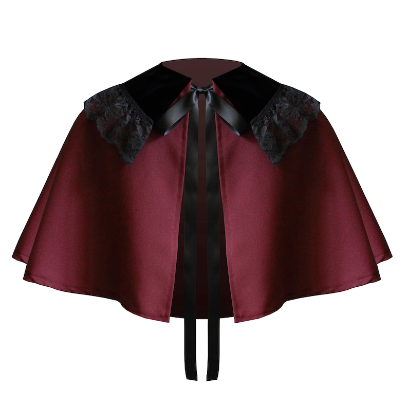 May include: A deep burgundy capelet featuring a wide black velvet collar adorned with black lace trim along the edges. The garment is secured at the neckline with a thin black satin ribbon tie that hangs down the front, creating a structured silhouette.