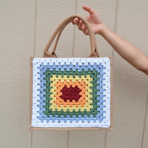 May include: A square tote bag with a colorful crochet design. The crochet pattern features a central red square surrounded by layers of orange, yellow, green, and blue, framed by a white border. The bag has tan handles and a burlap-like body.