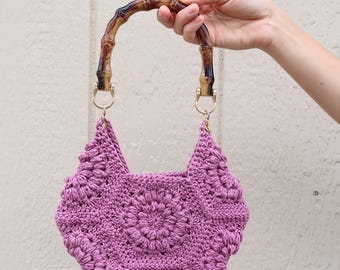 May include: A handcrafted, crochet bag in a soft purple hue. The bag features a floral pattern and is held by a bamboo handle with gold-colored metal hardware. The bag's design is unique and eye-catching.