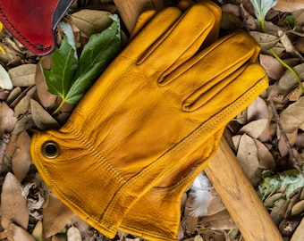 May include: A pair of yellow leather work gloves with a brown leather axe handle and a red leather axe sheath in the background. The gloves are lying on a bed of brown leaves.