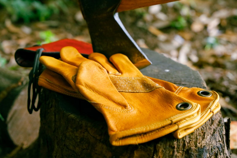 May include: A pair of yellow leather work gloves with reinforced stitching and metal eyelets. The gloves are lying on a wooden stump next to an axe.