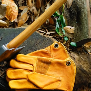 May include: A pair of yellow leather work gloves with a brown leather patch on the back of the hand. The gloves are lying on a tree stump with a wooden handled axe beside them.