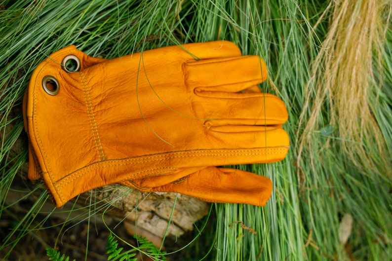 May include: A pair of yellow leather work gloves with a stitched seam detail. The gloves have two metal eyelets on the wrist.