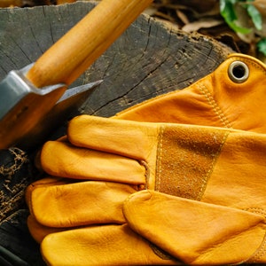 May include: A pair of brown leather work gloves with a reinforced palm and a metal rivet on the wrist strap. The gloves are lying on a wooden surface next to an axe.