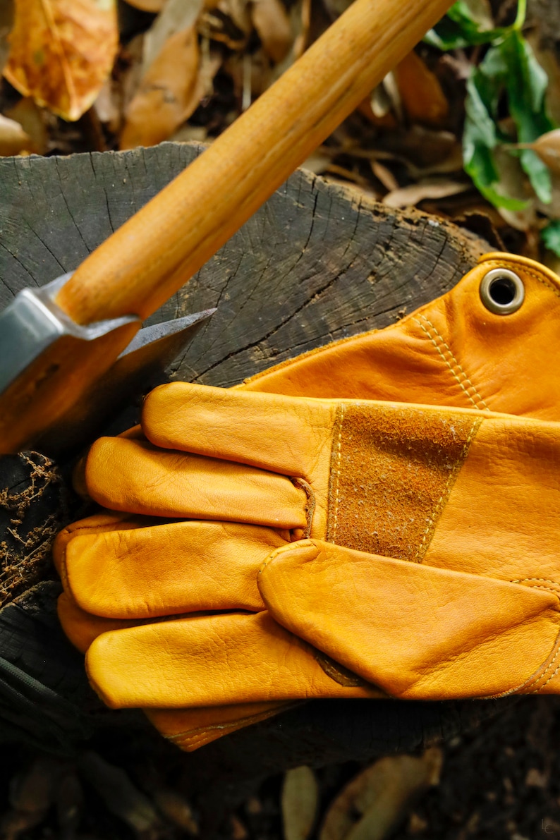 May include: A pair of brown leather work gloves with a reinforced palm and a metal rivet on the wrist strap. The gloves are lying on a wooden surface next to an axe.