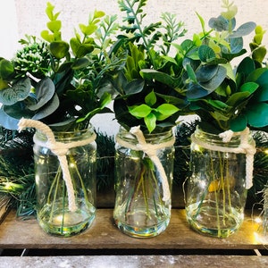 May include: Three glass jars with twine tied around the top, each filled with a bouquet of green leafy plants. The jars are sitting on a wooden surface.