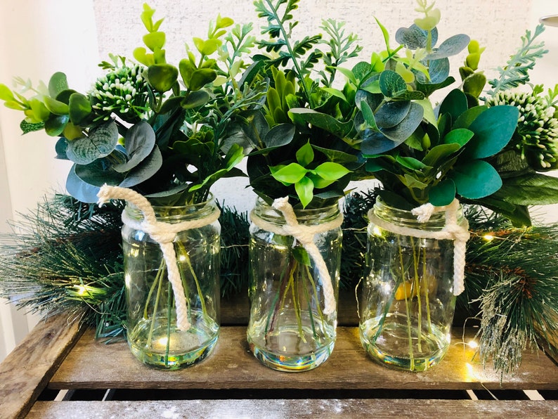 May include: Three glass jars with twine tied around the top, each filled with a bouquet of green leafy plants. The jars are sitting on a wooden surface.
