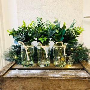 May include: Three glass jars with green leafy plants and twine tied around the top of each jar. The jars are sitting on a wooden crate with a white background.