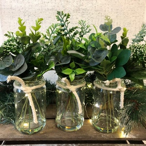 May include: Three clear glass jars with white twine tied around the top, each filled with a bouquet of green leafy plants.