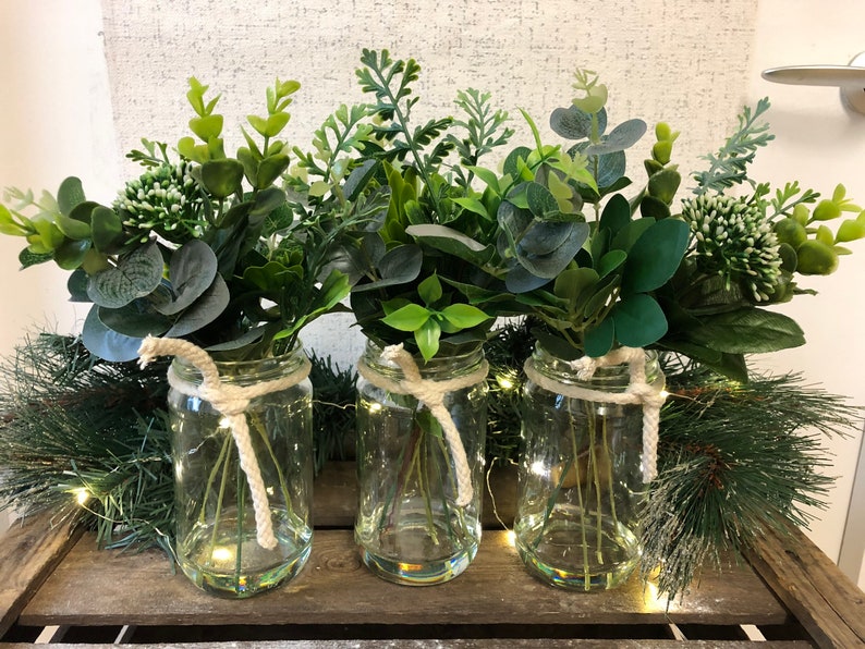 May include: Three clear glass jars with white twine tied around the top, each filled with a bouquet of green leafy plants.