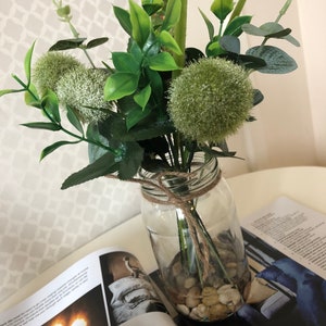 May include: A clear glass jar filled with small white stones and a bouquet of green artificial flowers. The flowers include green pom poms and long green leaves.