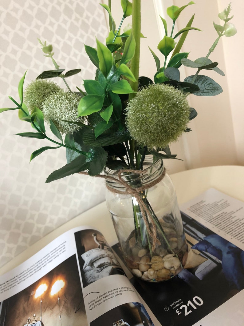 May include: A clear glass jar filled with small white stones and a bouquet of green artificial flowers. The flowers include green pom poms and long green leaves.
