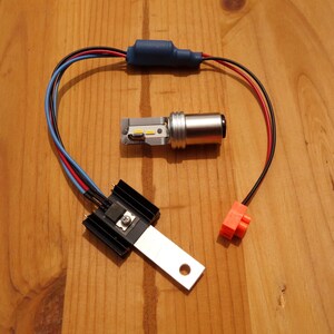 Può includere: Un assortimento di componenti per luci a LED per auto, tra cui un dissipatore di calore argento e nero, una lampadina a LED argento e vari fili con connettori. I componenti sono disposti su una superficie di legno.