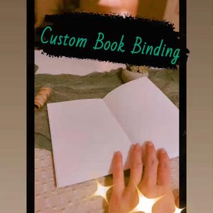 Puede incluir: Imagen de un libro abierto con páginas blancas en blanco, con el texto "Custom Book Binding" en fuente turquesa. El libro está sobre una superficie de color claro. Estallidos de estrellas decorativas en la esquina inferior derecha.