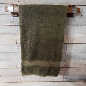 May include: A rustic wooden towel rack with a dark olive green towel hanging from it. The towel has a lighter green stripe at the bottom. The rack is mounted on a white wooden plank wall.