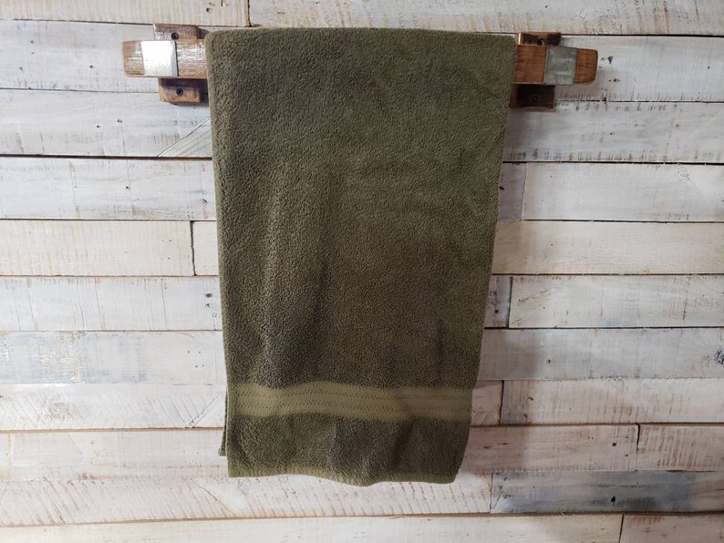 May include: A rustic wooden towel rack with a dark olive green towel hanging from it. The towel has a lighter green stripe at the bottom. The rack is mounted on a white wooden plank wall.