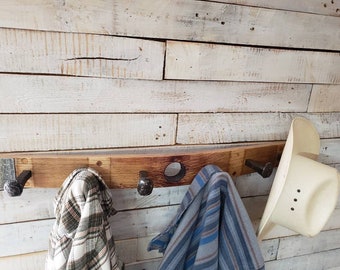 May include: A rustic wooden coat rack made from a repurposed barrel stave, featuring four black metal hooks. A plaid shirt, a blue striped shirt, and a white cowboy hat hang from the rack. The rack is mounted on a white-painted wooden wall.
