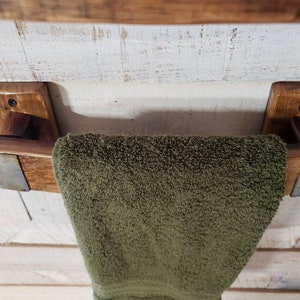 May include: A rustic wooden towel rack with a dark stain, featuring a green towel. The rack is mounted on a white, distressed wooden wall. The towel rack has metal accents.