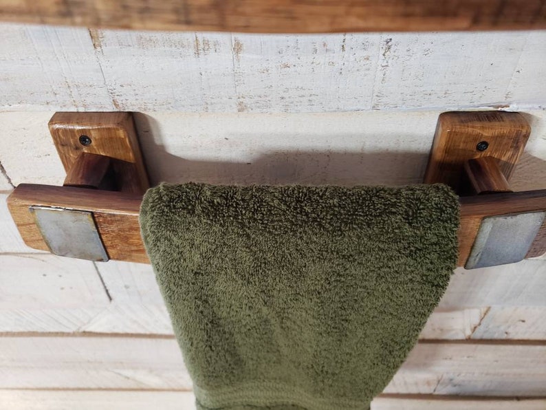 May include: A rustic wooden towel rack with a dark stain, featuring a green towel. The rack is mounted on a white, distressed wooden wall. The towel rack has metal accents.