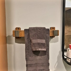 May include: A wooden towel rack with a gray towel hanging on it. The rack is made of light brown wood with gray metal accents. The towel is folded and has a textured pattern on the border. The rack is mounted on a white wall.