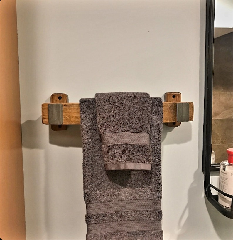 May include: A wooden towel rack with a gray towel hanging on it. The rack is made of light brown wood with gray metal accents. The towel is folded and has a textured pattern on the border. The rack is mounted on a white wall.