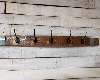 May include: A rustic wooden coat rack with five black metal hooks. The rack is made from a piece of reclaimed wood with a natural wood grain finish. The hooks are evenly spaced along the length of the rack, which is mounted on a white wooden plank wall.
