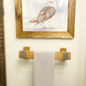 May include: A framed watercolor painting of a barn owl hangs above a wooden towel rack. The towel rack is made of light-colored wood with metal accents, holding a gray and white striped towel. The artwork and rack are mounted on a white wall.