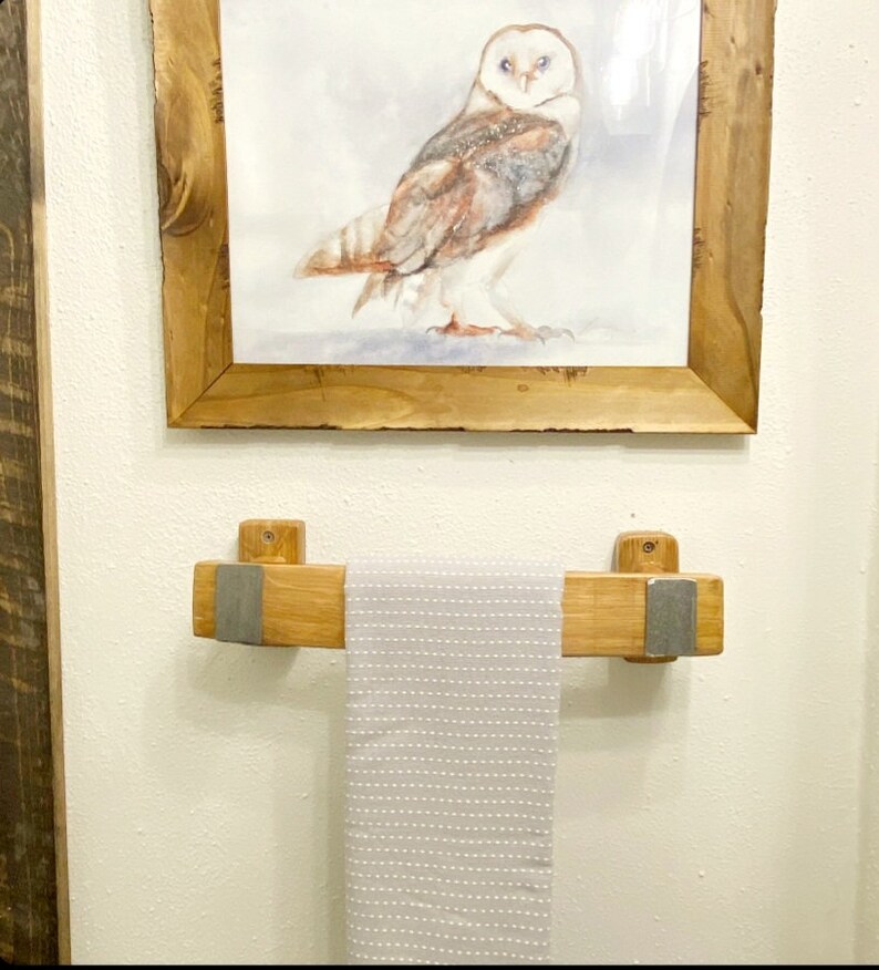 May include: A framed watercolor painting of a barn owl hangs above a wooden towel rack. The towel rack is made of light-colored wood with metal accents, holding a gray and white striped towel. The artwork and rack are mounted on a white wall.