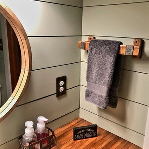 May include: A bathroom scene featuring a wooden towel rack with a gray towel. The rack is mounted on a light green wall. A round mirror and a soap dispenser are also visible. A sign that says "WASH YOUR HANDS" is on the counter.