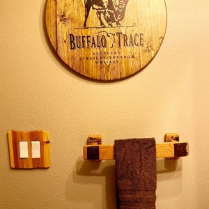 May include: A wooden wall decor piece with the text "BUFFALO TRACE" and a bison illustration. Below, a wooden towel rack holds a brown towel. A wooden light switch cover is to the left.