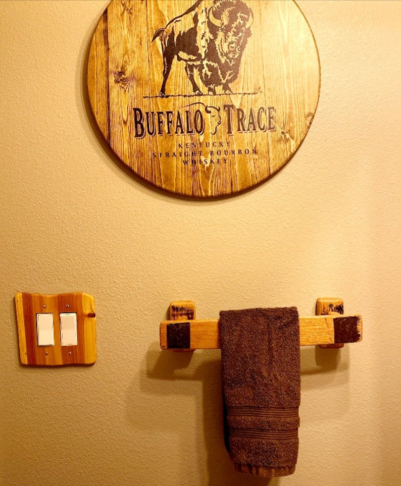 May include: A wooden wall decor piece with the text "BUFFALO TRACE" and a bison illustration. Below, a wooden towel rack holds a brown towel. A wooden light switch cover is to the left.