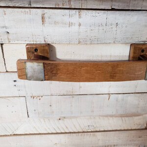 May include: A handcrafted wooden towel rack made from reclaimed oak barrel staves. The rack features a curved design with metal accents and is mounted on two rectangular wooden blocks. The wood has a natural, warm brown tone.
